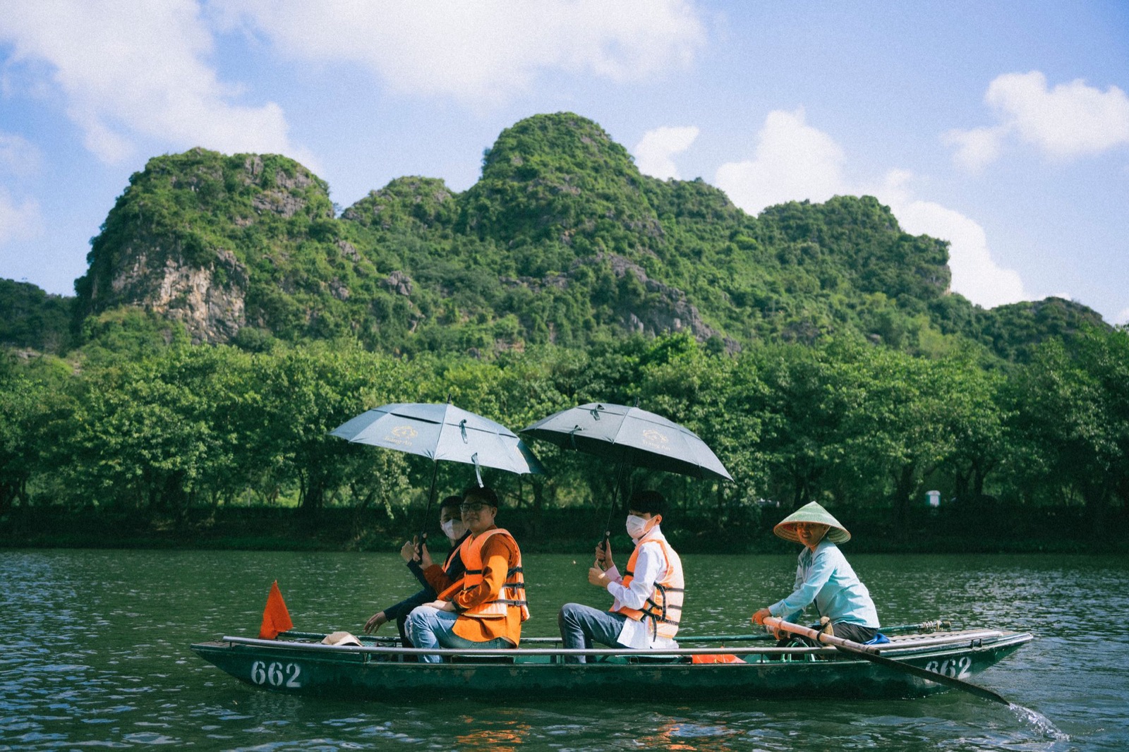 A rower in a conical hat steering a small boat under limestone cliffs in Ninh Bình